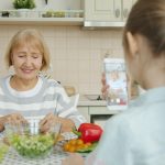 Cheerful elderly woman is posing for smartphone camera and cooking salad while child is taking picture with modern device. Family, photograph and people concept.