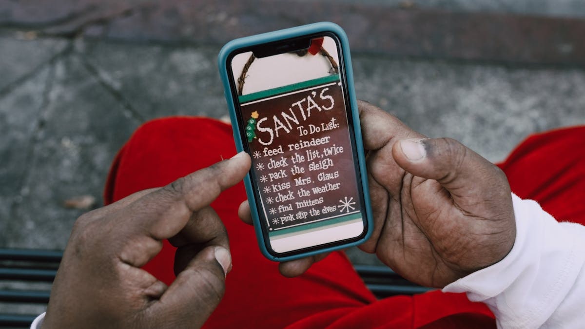 Close-up of hands holding a smartphone displaying Santa's whimsical to-do list outdoors.